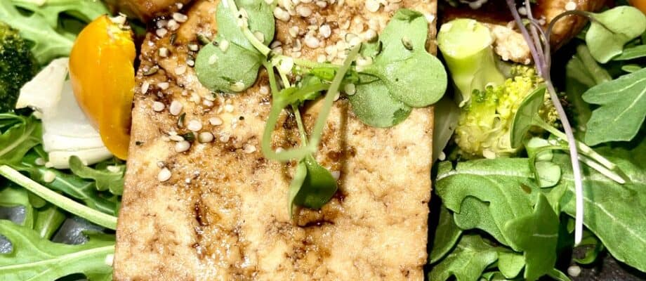 Grilled tofu salad with microgreens, mixed greens, cherry tomatoes, and seeds on a black plate.