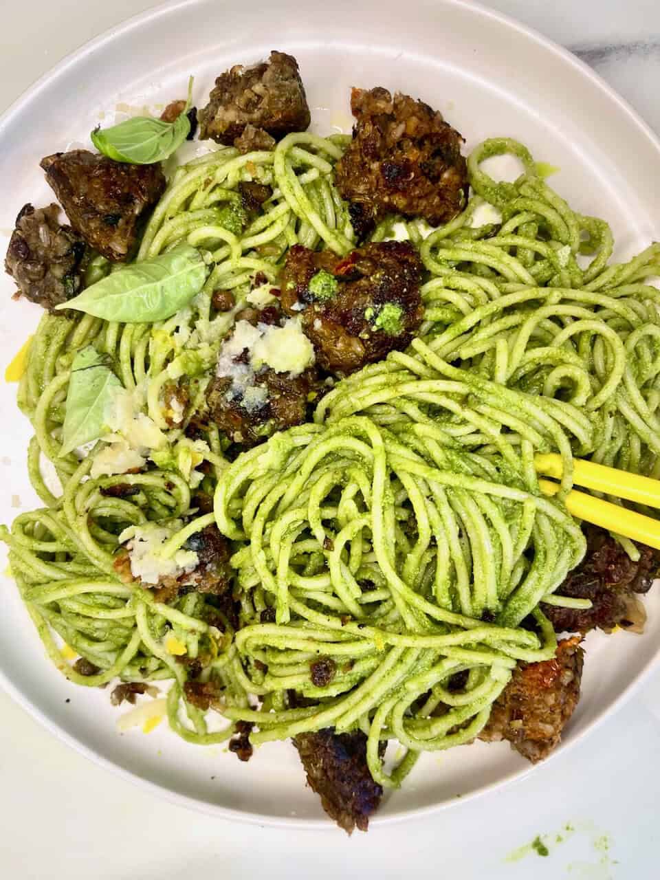 Green pasta with meatballs, basil, and grated cheese on a white plate.