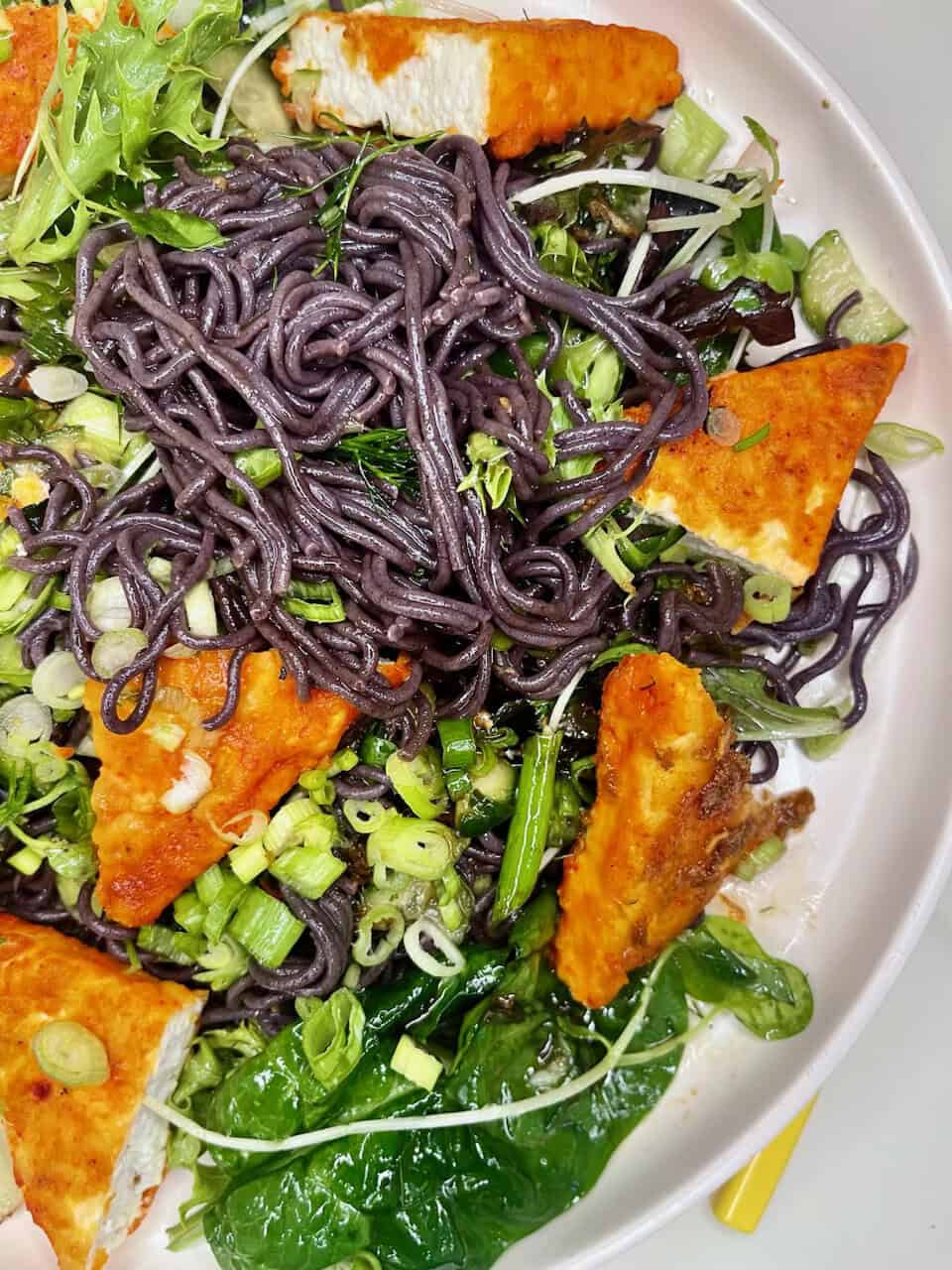 Dark purple black bean noodles with fresh greens and crispy tofu triangles, topped with chopped green onions and herbs.