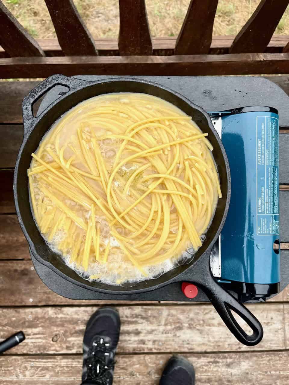 Cooking pasta in a black skillet on outdoor stove, yellow noodles boiling in water, rustic wooden deck background.