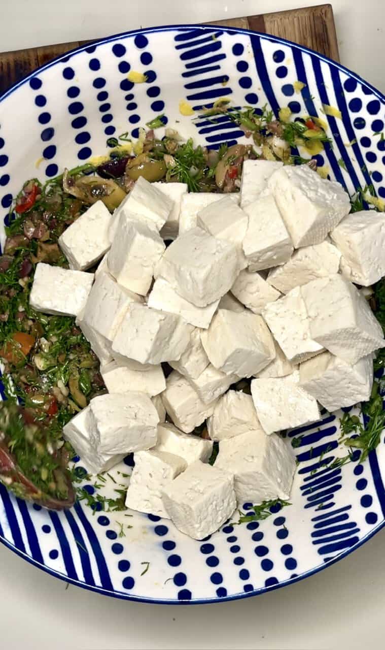 Fresh feta cheese cubes on a vibrant, patterned blue and white ceramic plate with mixed salad greens and vegetables in the background.