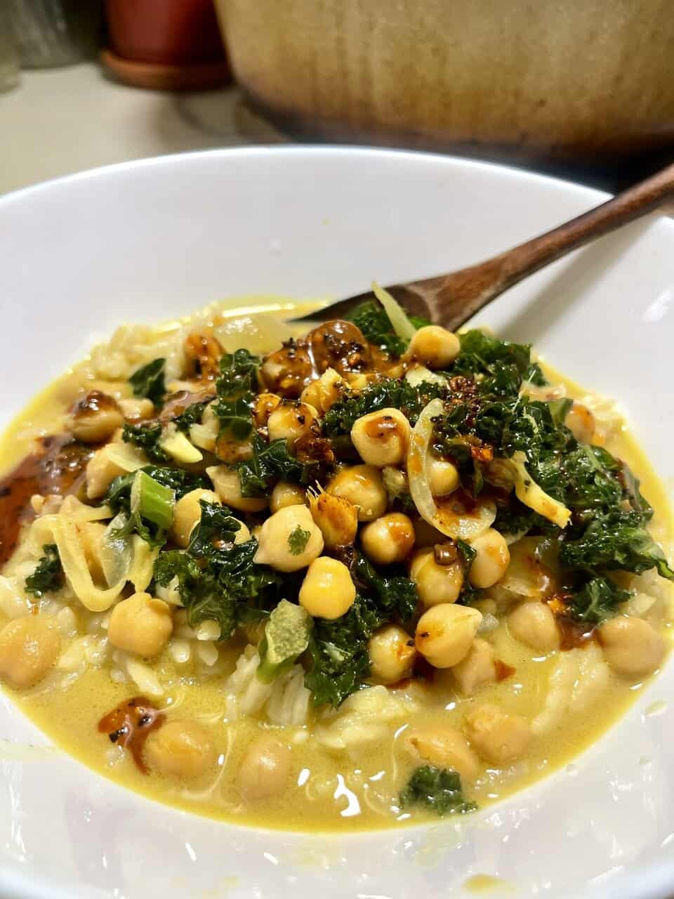 Chickpea and kale soup served in a white bowl with a wooden spoon, featuring creamy broth and fresh greens.
