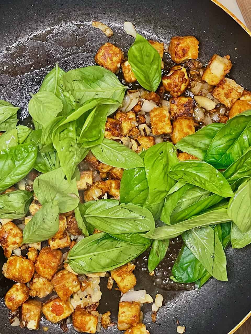 Basil and tofu being cooked in a skillet with onions and garlic.
