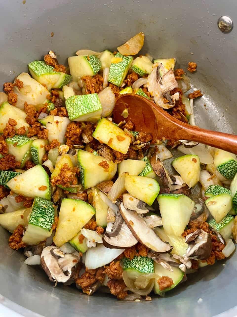Fresh zucchini, mushrooms, onions, and ground meat being sautéed in a cooking pot.