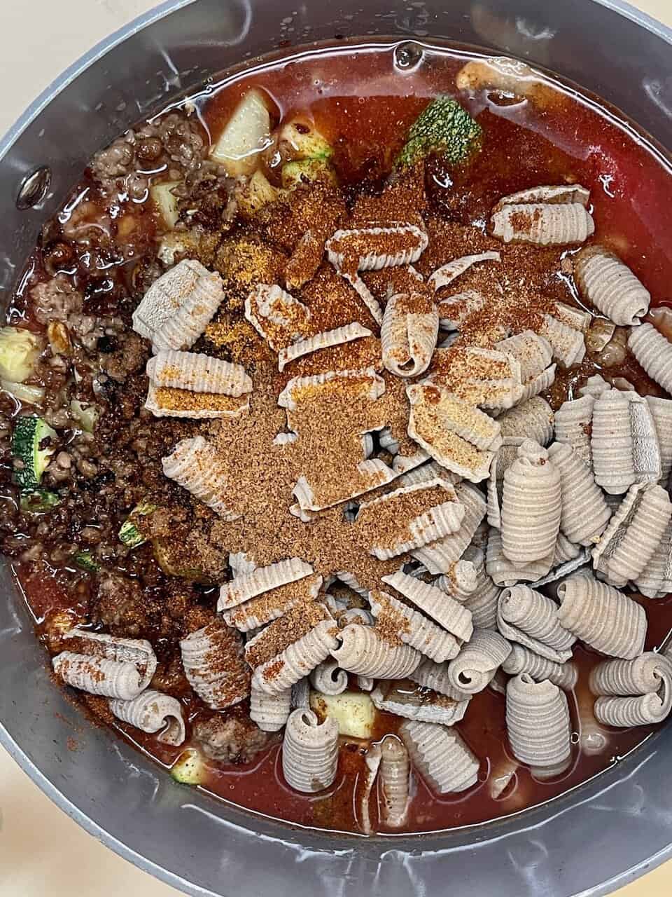 Beef and vegetable soup with dry pasta, spices, and broth in a cooking pot.