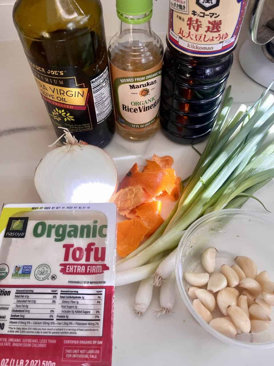 Bright soy sauce bottles, garlic cloves, green onions, tofu, rice vinegar, and orange peels on a kitchen countertop.