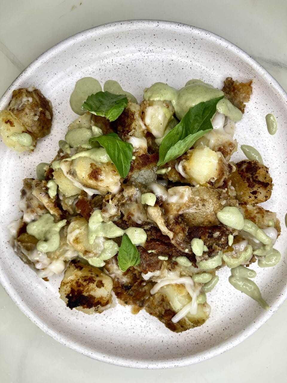 Crispy smashed potatoes with vegan herb sauce, fresh basil, and melted cheese on a white ceramic plate.