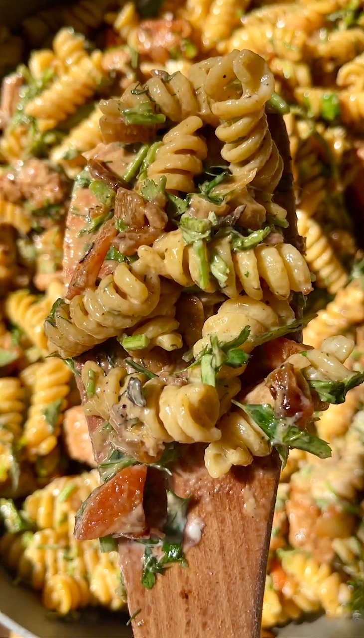 Close-up of pasta salad with herbs, bacon, and vegetables on a wooden spoon.