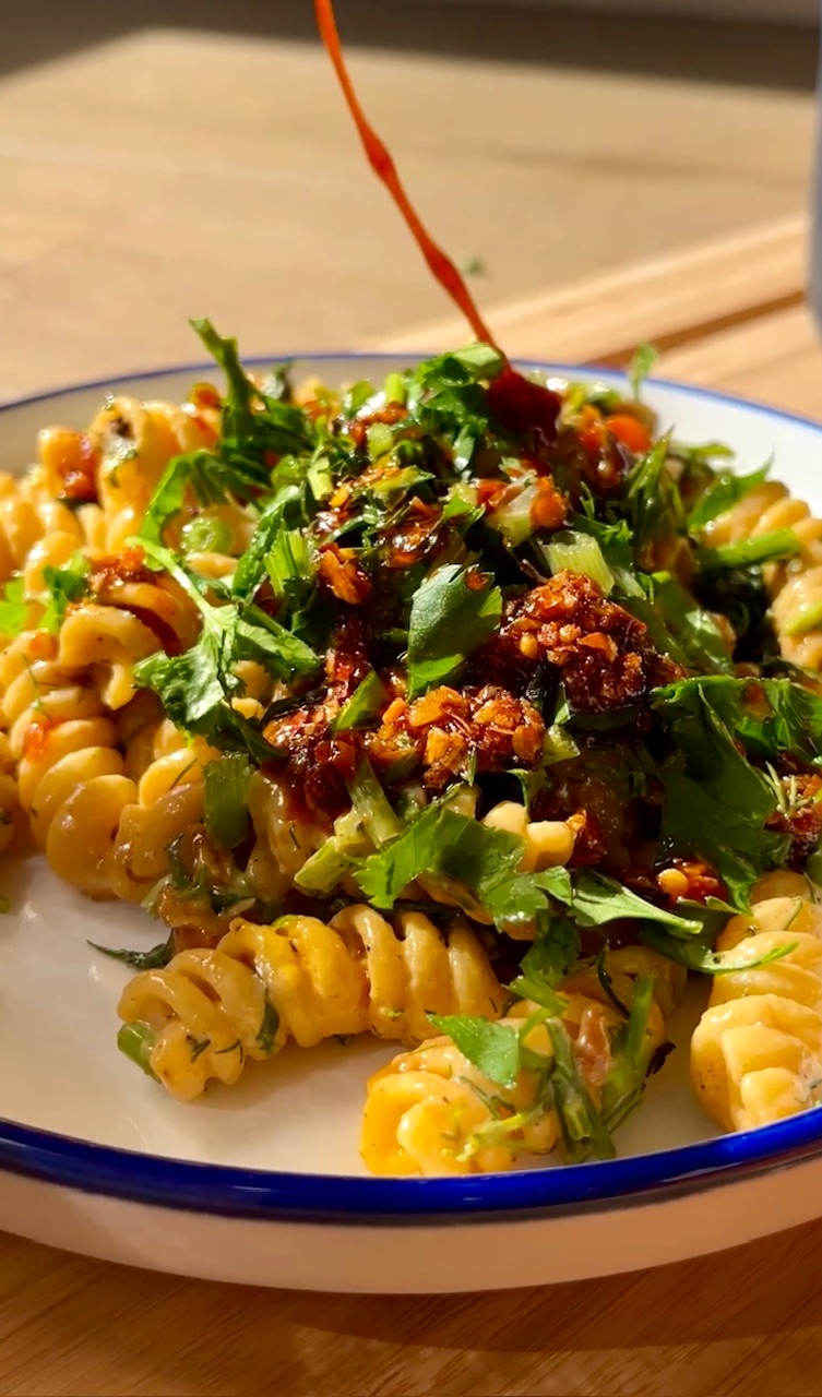 Spicy pasta with fresh herbs, chili sauce, and crunchy toppings on a white plate.