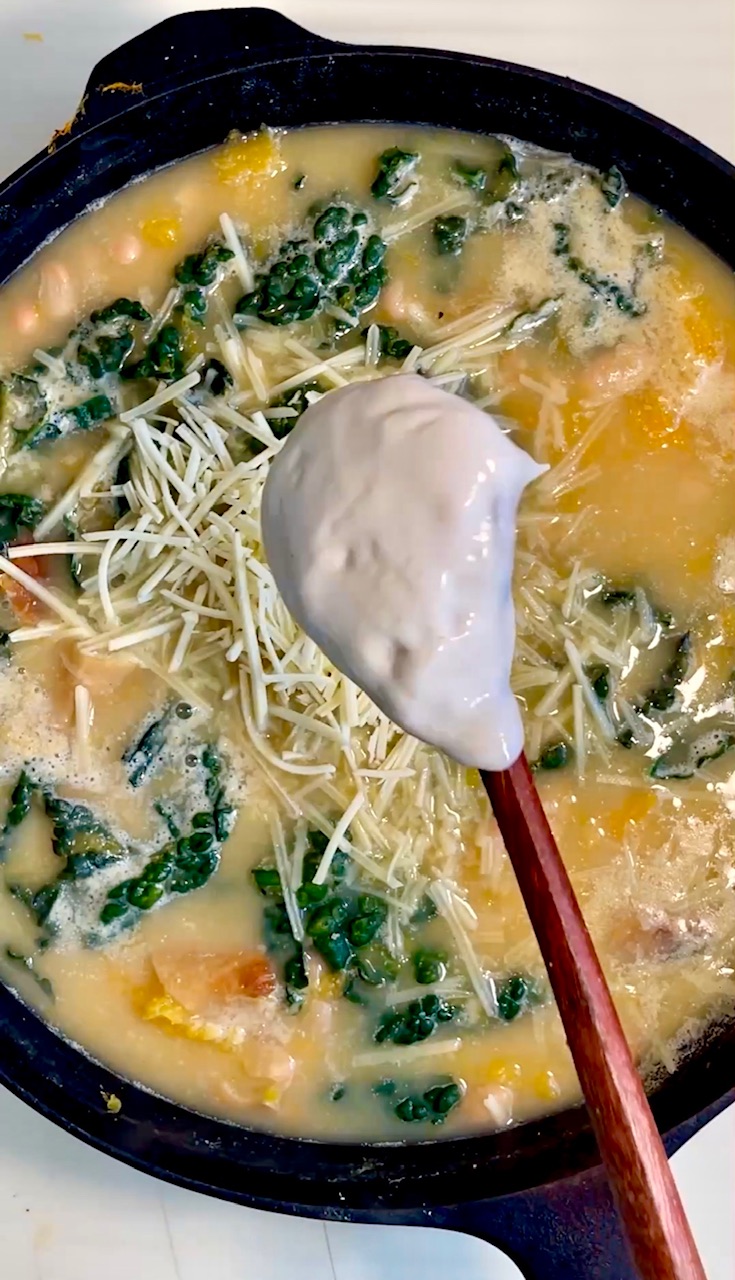 Creamy vegan stew with butternut squash, white beans, kale, and shredded cheese crumbles in a pot.